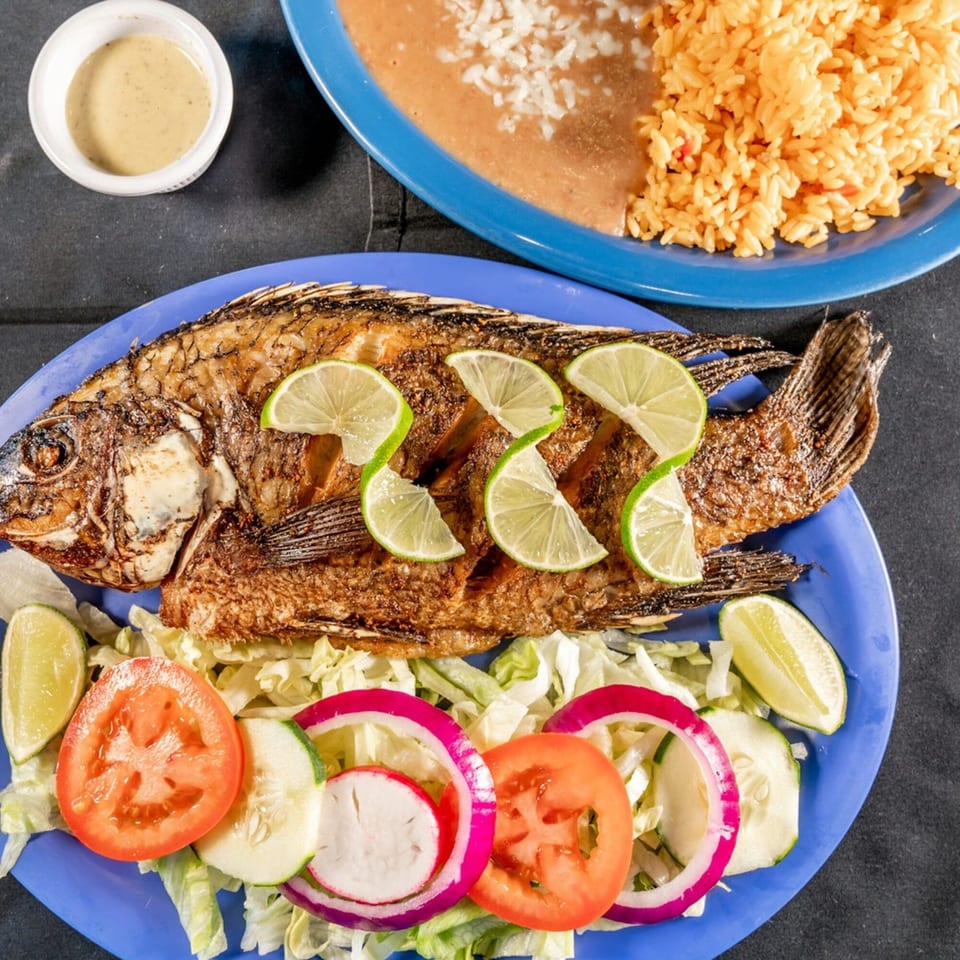 Fried whole fish with lime slices, lettuce, tomato, cucumber, and red onion, served with rice and beans.