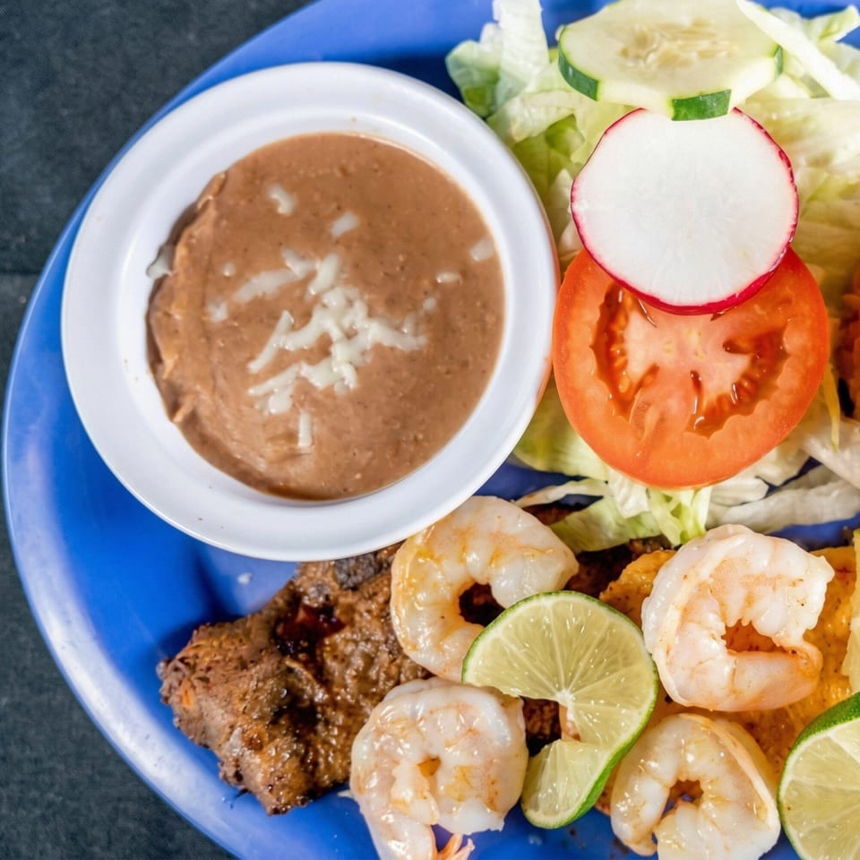 A blue plate with refried beans, salad, grilled meat, shrimp, and lime wedges.